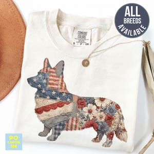 Puede incluir: Camiseta color crema con un diseño de Corgi en patchwork. La cabeza y las orejas del Corgi están estampadas con la bandera americana, mientras que el cuerpo presenta patrones florales y de patchwork en rojo, blanco y azul. El texto "ALL BREEDS AVAILABLE" está en un círculo azul marino.