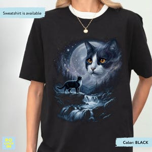 May include: A black t-shirt featuring a graphic print of a black and white cat standing on a rock near a waterfall, with a large cat face appearing in the starry night sky above. The text "Sweatshirt is available" is printed in the top left corner.
