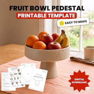 Puede incluir: Un pedestal de frutero beige lleno de naranjas, manzanas y plátanos. El texto "FRUIT BOWL PEDESTAL PRINTABLE TEMPLATE" está en la parte superior. Las palabras "EASY TO MAKE" están en un cuadro blanco con una carita sonriente. Varias plantillas imprimibles están frente al cuenco.