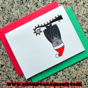 May include: A white greeting card featuring a cartoon bat wearing a Santa hat, hanging upside down from a branch decorated with Christmas lights. The card is on top of a green and red envelope. The website address www.oncupcakemoon.com is at the bottom.