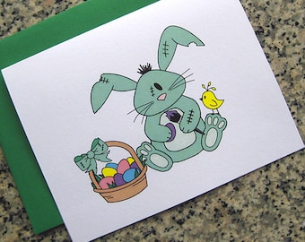 May include: A green stitched bunny rabbit holds a purple egg and a paintbrush. A yellow bird sits on a branch above the bunny. The bunny is next to a basket of colorful Easter eggs.