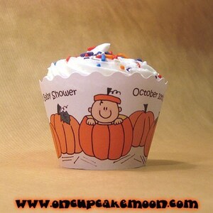 Puede incluir: Un cupcake blanco con chispas de color naranja, azul y blanco en un envoltorio de cupcake blanco con un dise&ntilde;o de calabaza y beb&eacute;. El envoltorio dice "Baby Shower" y "Octubre 2012".