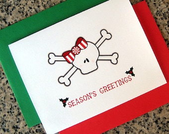 May include: A white card with a red and white candy cane skull and crossbones design. The card has the text "Season's Greetings" in red letters and a small green holly sprig. The card is layered on top of a green card and a red card.