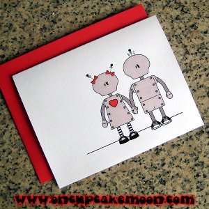 May include: A greeting card with a red envelope features a couple of robots holding hands. The robots are gray with red hearts and black details. The text on the card reads "www.oncupcakemoon.com".