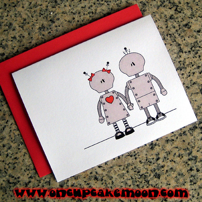 May include: A greeting card with a red envelope features a couple of robots holding hands. The robots are gray with red hearts and black details. The text on the card reads "www.oncupcakemoon.com".