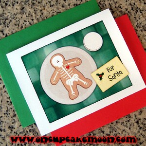 May include: A white card with a green background and a gingerbread man with a skeleton inside. The gingerbread man has a red heart on its chest. The card says "For Santa" on a small yellow tag with a holly sprig.