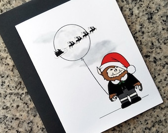May include: A black and white illustration of a werewolf wearing a Santa hat and holding a balloon with Santa Claus and reindeer flying in the moon. The text "www.oncupcakemoon.com" is at the bottom of the card.