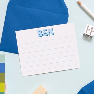 May include: A white card with blue lines and the name "BEN" in blue letters. The card is on a blue envelope and a light blue surface. There is a pencil sharpener, a pencil, and a small eraser on the surface.