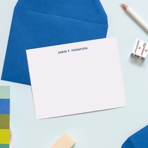 May include: A white card with a blue border and the text "JAMES P. THOMPSON" printed in blue at the top. The card is lying on a light blue surface with a blue envelope, a pencil, a pencil sharpener, and a small card with colorful text.