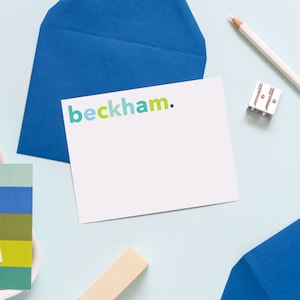 May include: A white card with the text "beckham*" in colorful letters on a blue background. The card is on a light blue surface with a white pencil, a pencil sharpener, and a white card with colorful text.