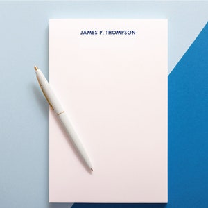 May include: A white notepad with a blue header that says "JAMES P. THOMPSON" on a light blue and blue background with a white pen on top of the notepad.