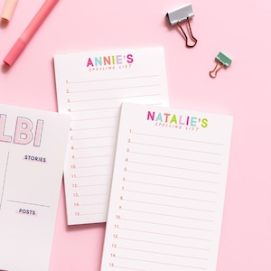 May include: Two white notepads with colorful text on the top. The top notepad says "ANNIE'S SPELLING LIST" and the bottom notepad says "NATALIE'S SPELLING LIST". Both notepads have numbered lines for writing.