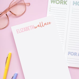 May include: A white notepad with the text "ELIZABETHwallace" in pink and orange lettering. The notepad is on a pink surface with a pair of pink glasses and two pens, one yellow and one blue.