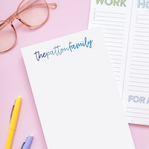 May include: A white notepad with the text "thepattonfamily" written in blue and a gradient of blue colors. The notepad is on a pink surface with two pens, one yellow and one purple, and a pair of pink glasses.