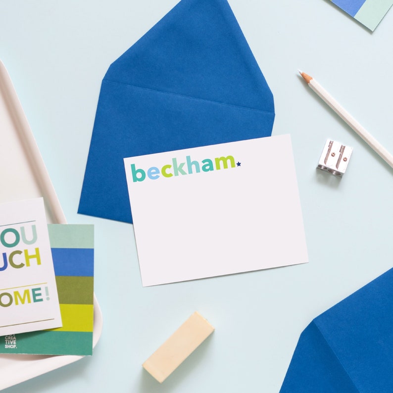 May include: A white card with the text "beckham*" in colorful letters on a blue background. The card is on a light blue surface with a white pencil, a pencil sharpener, and a white card with colorful text.