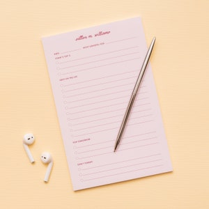 May include: A pink notepad with a silver pen and white earbuds. The notepad has a pink and white striped design and the text "outton m. williamo" at the top. The notepad has sections for "MOST GRATEFUL FOR", "TODAY'S TOP 3", "NEXT ON THE LIST", "FOR TOMORROW", and "DON'T FORGET".