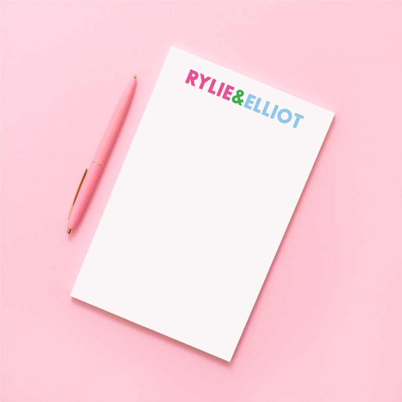 Puede incluir: Un bloc de notas blanco con el texto "RYLIE & ELLIOT" en letras de colores. Un bol&iacute;grafo rosa est&aacute; sobre el bloc de notas. El bloc de notas y el bol&iacute;grafo est&aacute;n sobre un fondo rosa.