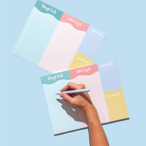 May include: Two rectangular paper planners with pastel color-blocked sections labeled Things to do, What to get, What to call, and Email back in white script. One planner is blank, while the other is being written on with a light blue pen.