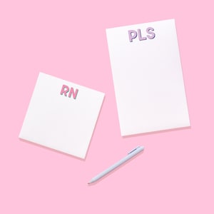 May include: Two white paper notepads and a light blue pen. The square notepad features the pink letters RN, while the rectangular notepad displays the purple letters PLS. The items are arranged on a solid light pink background surface.
