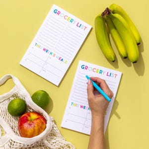 May include: Two white grocery list notepad pads with rainbow lettering. The top pad has the text "Grocery List" and "This Week's Meals". The bottom pad has the text "Grocery List" and "This Week". A hand is writing on the bottom pad with a blue pen. There are bananas, limes, and an apple in the background.