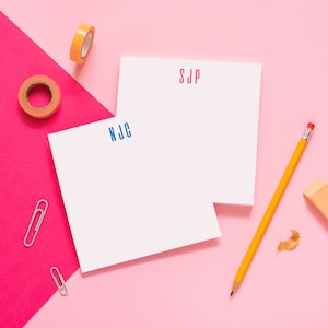 May include: Two white notepads with blue and pink initials "NJC" and "SJP" on a pink and red background. A yellow pencil, a pencil sharpener, and two silver paper clips are also on the surface.