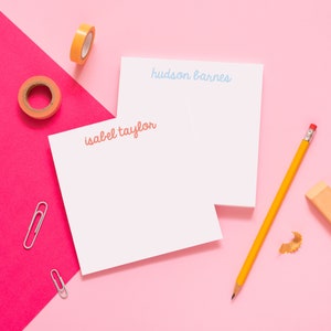 May include: Two white notepads with different names written on them in colorful script. The top notepad says "hudson barnes" in light blue and the bottom notepad says "isabel taylor" in orange. The notepads are on a pink surface with a yellow pencil, a pink eraser, and two silver paper clips.