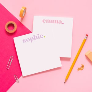 May include: Two white notepads with pink and purple text. The top notepad says "emma." and the bottom notepad says "sophie.". A yellow pencil, a pink eraser, and two silver paper clips are on a pink surface.