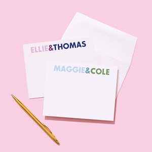May include: Two white note cards with colorful text. The top card says "ELLIE & THOMAS" in purple and blue. The bottom card says "MAGGIE & COLE" in blue, light blue, and green. A gold pen is lying on a pink surface.