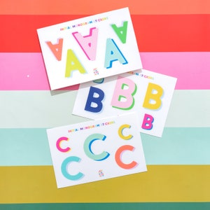May include: Three sheets of colorful initial monogram stickers. Each sheet features a different letter of the alphabet: A, B, and C. The letters are in various colors, including pink, blue, green, yellow, and orange. The sheets are labeled "Initial Monogram Stickers".