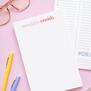 May include: A white notepad with the text "maggiesmith." printed in pink and orange letters. The notepad is on a pink surface with two pens, one yellow and one blue, and a pair of pink glasses. A white notepad with a list titled "WORK" and another list titled "HOME" is visible in the background.