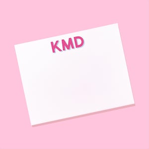 May include: A rectangular white notepad featuring the letters KMD printed in a bold, dark pink font with a light blue shadow effect. The notepad is positioned against a solid, light pink background, providing a clean and simple visual presentation.