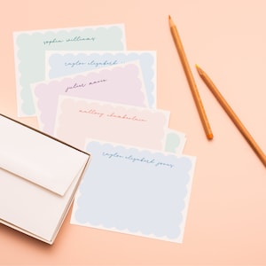 May include: A set of five pastel colored stationery note cards with scalloped edges. Each card has a different name written in cursive on the top. The cards are arranged in a stack with the top card being a light blue color. The cards are on a pink surface with two wooden pencils.