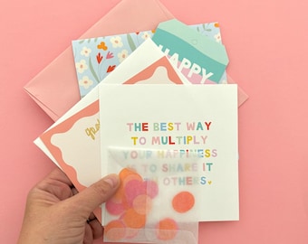 Könnte beinhalten: Ein Stapel Grußkarten und Umschläge, darunter eine weiße Karte mit dem Text THE BEST WAY TO MULTIPLY YOUR HAPPINESS IS TO SHARE IT WITH OTHERS in bunten Buchstaben, sowie ein kleiner, durchscheinender Umschlag gefüllt mit orangefarbenem Konfetti.