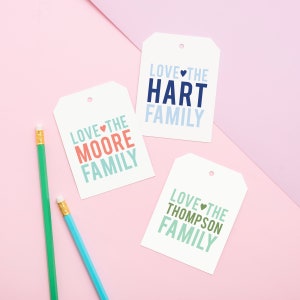May include: Three white gift tags with a hole punched in the top. Each tag has the text "Love the" followed by a family name in colorful letters. The names are Moore, Hart, and Thompson. The tags are on a pink background.