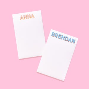 May include: Two rectangular white paper notepads. The top notepad features the name ANNA in orange block letters with a subtle shadow. The bottom notepad features the name BRENDAN in light blue block letters with a subtle shadow effect.