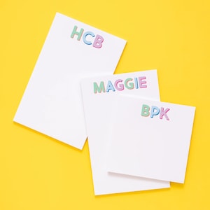 May include: Three white notepad sets with colorful letters spelling out the names "HCB", "MAGGIE", and "BKP". The notepads are on a yellow background.