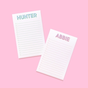 May include: Two rectangular white notepads with lined paper and circular checkboxes. One notepad features the name HUNTER in light blue letters with green outlines, while the other displays the name ABBIE in pink textured letters.