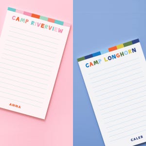 May include: Two white rectangular notepads with horizontal blue lines. The left notepad features the text CAMP RIVERVIEW in multicolored letters with a striped header and the name ANNA at the bottom. The right notepad displays CAMP LONGHORN in multicolored letters with a striped header and the name CALEB at the bottom.