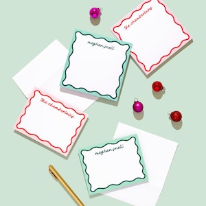 May include: An overhead shot of stationery items, including notecards and envelopes, arranged on a light green surface. The notecards have scalloped edges and feature names in script. Christmas ornaments and a gold pen are scattered around the cards.