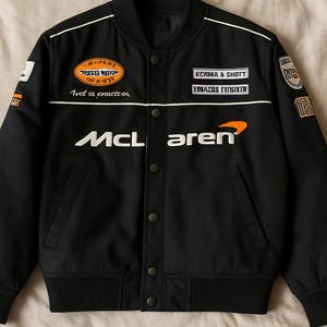 Può includere: Giubbotto bomber nero con dettagli bianchi e toppe arancioni e bianche. La giacca ha una chiusura a zip e un colletto alto. Le toppe includono le parole "Red Bull", "Ecma & Schott", "Edjazus Genisitin" e "McLaren".