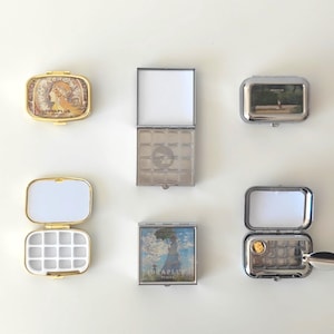 Peut inclure: Plusieurs petits contenants métalliques rectangulaires avec des couvercles à charnière. Certains sont dorés, d'autres argentés. L'un présente un portrait, un autre un paysage, et l'un est ouvert avec une grille de petits compartiments.