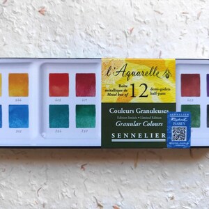 Pode incluir: Uma caixa metálica Sennelier contendo 12 meios godets de aguarela em cores granulares. O conjunto apresenta uma paleta de amarelo, laranja, vermelho, azul, verde e roxo com códigos de cor numerados. Um rótulo central diz l'Aquarelle 12 demi-godets.