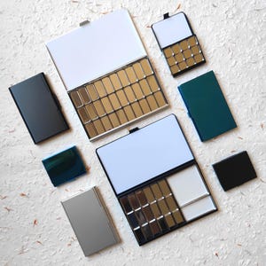 Può includere: Una collezione di scatole rettangolari in metallo per acquerelli, di varie dimensioni e colori, tra cui nero, argento, verde acqua e blu. Alcune scatole sono aperte, rivelando file di vaschette metalliche vuote e aree bianche per miscelare.