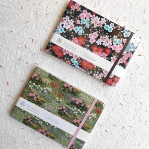 Op de afbeelding: Twee rechthoekige schetsboeken met elastieken banden. Het bovenste schetsboek heeft een zwarte kaft met roze, rode en blauwe kersenbloesempatronen. Het onderste schetsboek heeft een groene kaft met een veelkleurige bloementuinprint en witte papieren banden.