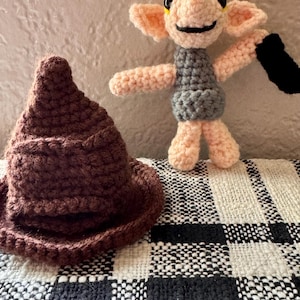 May include: A handmade crochet Sorting Hat in brown and a Dobby doll with a gray tunic, holding a black sock. The doll has pink arms, legs, and ears. The background is a textured wall and a black and white checkered blanket.