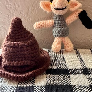 May include: A handmade crochet Sorting Hat in brown and a Dobby doll with a grey tunic, holding a black sock. The doll has pink arms, legs, and ears. The background is a textured wall and a black and white checkered blanket.