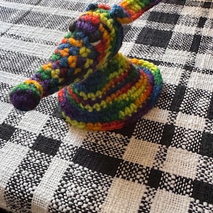 May include: A handmade crochet creature featuring a vibrant rainbow-colored yarn pattern throughout its body. The figure has a long, pointed snout, small dark eyes, and a wide, circular base that allows it to stand upright on a flat surface.