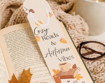May include: A beige bookmark with the text "Cozy Reads & Autumn Vibes" and fall-themed illustrations, including a coffee cup, books, and leaves. The bookmark is placed in an open book with a fall leaf.