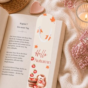 Puede incluir: Marcapáginas beige con la frase "Hello Autumn" e ilustraciones de manzanas rojas, hojas naranjas y un erizo. El marcapáginas está en un libro abierto con texto en alemán.