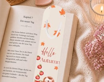 May include: A beige bookmark with the text "Hello Autumn" and illustrations of red apples, orange leaves, and a cute hedgehog. The bookmark is placed in an open book with German text.